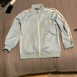 Ljusblå Palm Angels track jacket - Palm Angels ljusblå track jacket med vita och svarta ränder längs ärmarna. Jackan har ståkrage, hel dragkedja framtill och två fickor med dragkedja. Materialet är syntet och jackan har en sportig, loose passform. Ikonisk Palm Angels-logga på bröstet.