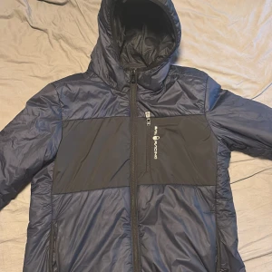 Svart-blå pufferjacka Sail Racing - Säljer en stilren pufferjacka från Sail Racing i svart och mörkblått. Jackan har huva, två dragkedjor framtill och logga på bröstet. Materialet är syntet och polyester, perfekt för kalla dagar. Jackan har lång ärm och en clean, sportig look.