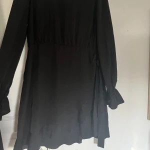 Svart långärmad kortklänning med knyt - En svart klänning med långa ärmar och rund halsringning. Klänningen har knytband vid midjan. Tunt, mjukt tyg som ger ett snyggt fall. 