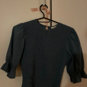 Mörkblå toppmed puffärm från NA-KD - Snygg mörkblå topp  från NA-KD med puffiga trekvartsärmar och volang vid ärmslut. Smockad överdel ger en cool och bekväm passform. Perfekt för dig som gillar stilrena detaljer och vill sticka ut med en unik ärmdesign.