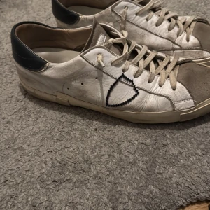 Vita sneakers från Phillipe model  - Säljer ett par vita sneakers i skinn med svarta detaljer på hälen och en broderad svart triangel på sidan. Skorna har snörning och en platt sula i gummi. Perfekta för dig som gillar en clean men ändå unik look. Skorna är lite slitna vid sulan men det går enkelt att fixa med lim! 