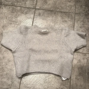 Ljusgrå croppad stickad tröja - Supermysig ljusgrå stickad tröja från Design by Si. Croppad modell med korta ärmar och rund halsringning. Perfekt att styla med höga jeans eller kjol för en chill och trendig look. Nypris ungefär 300