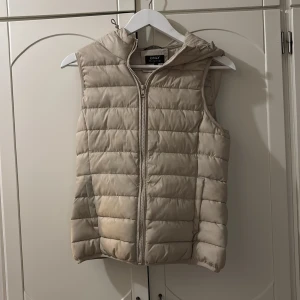 Beige dunväst med huva från Only - Snygg beige dunväst från Only med quiltad design och huva. Västen har dragkedja framtill och är perfekt för lager-på-lager-stil. Den är lätt vadderad och har en stilren look som passar till många outfits.