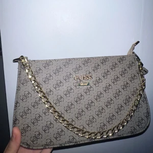Beige axelväska från Guess med kedja - Snygg beige axelväska från Guess med brunt logomönster över hela väskan. Väskan har en guldfärgad kedja som detalj och logotyp i guld framtill. Stilren form med dragkedja upptill och justerbar axelrem. Perfekt för dig som gillar trendiga accessoarer.
