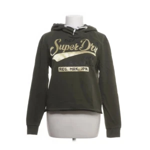 Olivgrön hoodie från Superdry - Snygg olivgrön hoodie från Superdry med tryck i beige och vitt framtill. Perfekt för en avslappnad och cool stil. Mjukt material och långa ärmar.