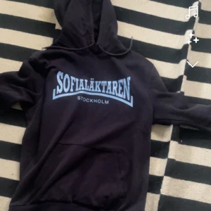 Svart hoodie Sofialäktaren Stockholm Djurgården  - Svart hoodie med ljusblå text 'Sofialäktaren Stockholm' tryckt på bröstet. Klassisk huva med snörning och stor magficka framtill. Perfekt för dig som vill visa support för Stockholm och Sofialäktaren.