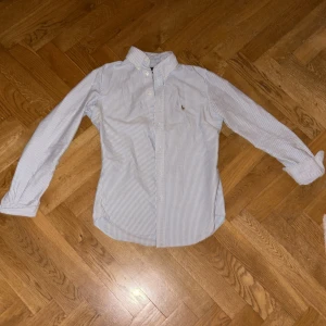 Randig skjorta från Ralph Lauren - Klassisk skjorta från Ralph Lauren i vitt och ljusblått med smala ränder. Skjortan har button down-krage, långa ärmar och broderad logga på bröstet. Tillverkad i mjuk bomull och har knäppning framtill.