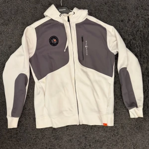 Vit och grå huvtröja från sail Racing - Snygg vit och grå huvtröja från Sail Racing med dragkedja och broderad logga på bröstet. Tröjan har långa ärmar och en praktisk bröstficka med dragkedja. Perfekt för en avslappnad stil. Storlek XL