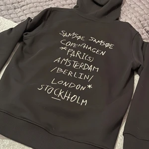 Svart hoodie från Samsøe Samsøe - Svart hoodie med coolt tryck av storstäder på ryggen och en vit logga på bröstet. Tröjan har huva med snörning, stor magficka och ribbade muddar. Perfekt för dig som gillar streetstyle och vill ha något som sticker ut. Använd fåtal gånger på grund av att den blev för stor och gick inte att lämna tillbaka. Ny pris på denna är 1700kr men säljer den för 700kr. Bara att skriva vid minsta fundering eller fråga mvh Liam.