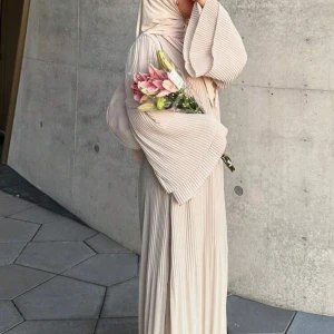 Plisserad beige långklänning - Superfin plisserad abaya/långklänning i beige med vida ärmar och snygga volangdetaljer vid ärmsluten. Klänningen har en flowy passform och är tillverkad i ett lätt, syntetiskt material som ger ett elegant fall. Perfekt för dig som gillar stilrena och trendiga looks. Passar storlekar S-XL och längder 160-170.