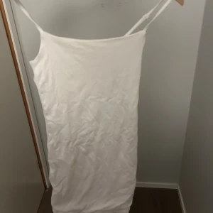 Vit ärmlös kortklänning med smala band - En enkel och stilren vit kortklänning med smala axelband. Klänningen är ärmlös och har en rak passform som sitter snyggt på kroppen. Perfekt för varma dagar eller att styla med accessoarer. Materialet känns mjukt och lätt.