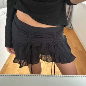 Svart volangkjol med snörning - Supersnygg svart kortkjol med volanger och snörningsdetaljer framtill. Perfekt för dig som gillar en trendig och feminin stil.