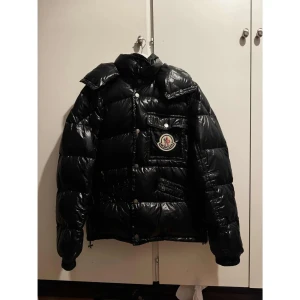 Svart pufferjacka från Moncler - Svart pufferjacka från Moncler med glansig finish och stor logopatch på bröstet. Jackan har huva, flera fickor med knappar och dragkedja framtill. Perfekt för kalla dagar och har en riktigt schysst streetstil. Storlek 4 vilket motsvarar L