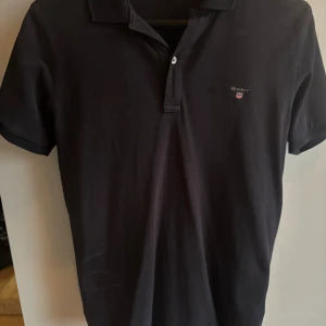 Svart pikétröja från GANT - Klassisk svart pikétröja från GANT med broderad logga på bröstet. Tröjan har korta ärmar, krage och knäppning framtill. Tillverkad i bomull för en skön och stilren känsla.