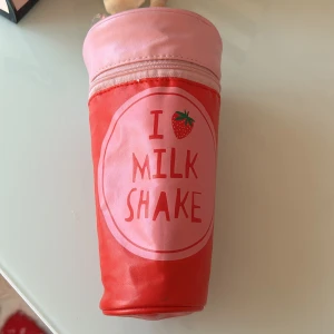 Rosa och röd milkshake-väska - Rolig och unik väska formad som en milkshake i rosa och rött med texten 'I love milk shake' och en jordgubbe på framsidan. Väskan har dragkedja upptill och är tillverkad i syntetmaterial. Perfekt för dig som vill sticka ut med en lekfull accessoar.