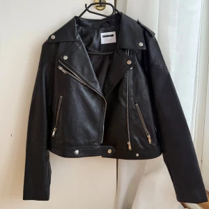 Svart skinnjacka från Noisy May - Snygg svart skinnjacka, köpt på Vero Moda med klassisk bikerstil. Jackan har silvriga dragkedjor, knappar och detaljer, samt bred krage och axelklaffar. Modellen är kortare och har långärmade ärmar. Perfekt för dig som vill ha en edgy look. Har knappt använt den.