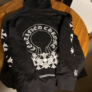Svart Chrome Hearts hoodie  - Svart hoodie från Chrome Hearts med stora vita tryck på ryggen och ärmarna. Hoodien har snörning vid huvan och en klassisk känguruficka framtill. Snygga detaljer med Chrome Hearts-logga och grafiska korsmönster längs ärmar och rygg. Perfekt för dig som gillar streetwear.