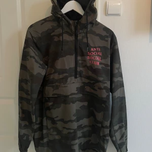 Camouflage vindjacka Anti Social Social Club - Säljer en snygg camouflage vindjacka från Anti Social Social Club med stor rosa logga på ryggen och mindre tryck på bröstet. Jackan har huva, half zip och stor ficka framtill. Perfekt för dig som gillar streetwear och vill sticka ut.