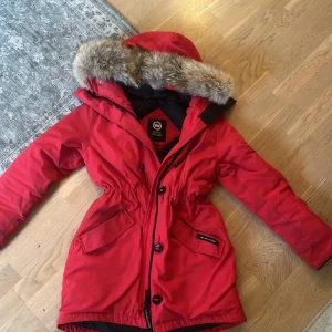 Canada goose - Snygg röd parkas frán Canada Goose med pälskantad huva och klassisk logga pả ärmen. Perfekt för kalla dagar med sin varma dunfyllning och justerbar midja for schysst passform. Lite skitig pa ärmar men gảr bort i tvätt, Liten i strl
