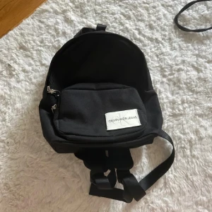 Calvin Klein väska - En svart ryggsäck från Calvin Klein Jeans med vit logotyp framtill. Väskan har justerbara axelband, ett stort huvudfack och en ytterficka med dragkedja. Fin men aldrig kommit till användning 