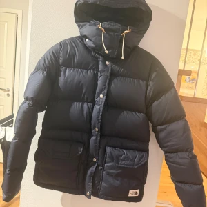 Mörkblå dunjacka från The North Face - Säljer en mörkblå dunjacka från The North Face med huva och snörning. Jackan har stora fickor framtill, knappar och dragkedja. Perfekt för kalla dagar och har en riktigt schysst puffer-look som är både trendig och bekväm.