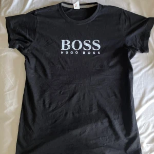 Svart BOSS t-shirt från Hugo Boss - Svart t-shirt från Hugo Boss med stor vit BOSS-logga på bröstet. Klassisk rund halsringning och korta ärmar. Tillverkad i mjuk bomull för en skön känsla och stilren look. Perfekt för dig som gillar enkel och snygg streetwear. Passform: slimfit/regularfit