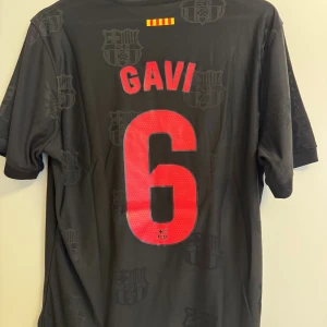 FC Barcelona Gavi #6 svart matchtröja - Snygg svart unik FC Barcelona fotbollströja från Nike med Gavi och nummer 6 på ryggen. Tröjan har klubbmärke, Nike-logga och Spotify-logga på bröstet samt diskreta klubbemblem i tyget. Tillverkad i lätt och ventilerande material, perfekt för match eller träning.