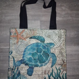 Shoppingväska med havssköldpadda - En stor shoppingväska med svart handtag gjord I ett tunnare material och ett färgglatt tryck av en blå havssköldpadda, sjöstjärna och havsväxter på framsidan även på baksidan. Bakgrunden har ett kartinspirerat mönster i beige och blå toner. Perfekt för att bära med sig allt man behöver. Mått ca 42x40cm 