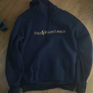 Marinblå hoodie Polo Ralph Lauren XL - Snygg marinblå hoodie från Polo Ralph Lauren.storlek Xl på lappen men jag har S/M i storlek och sitter bra på mig. Tröjan har gul broderad logga på luvan och stort Polo Ralph Lauren-tryck på bröstet. Klassisk huva med snörning och känguruficka framtill. Mjuk och skön bomullskvalitet. Ställ gärna frågor vid funderingar!😁
