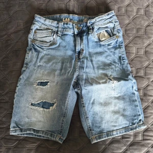 Blå slitna jeansshorts LAB Industries - Säljer ett par blå jeansshorts från LAB Industries med slitna detaljer och hål på benen. Klassisk femficksmodell med knapp och dragkedja. Perfekt för dig som gillar en avslappnad och trendig stil.