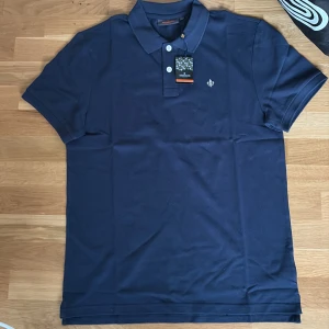 Marinblå piké från Morris Stockholm -  En snygg sprillans ny marinblå pikétröja från Morris Stockholm i storlek M. Klassisk design med krage, två knappar och diskret logga broderad på bröstet. Tillverkad i mjuk bomull som känns skön mot huden. Perfekt för dig som gillar stilrena och tidlösa plagg.