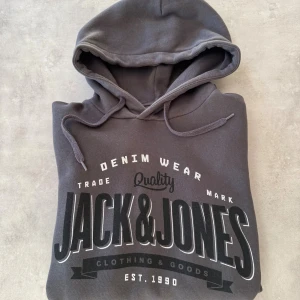 Mörkgrå hoodie Jack & Jones - Snygg mörkgrå hoodie från Jack & Jones med stor logga framtill. Tröjan har huva med snörning, magficka och mjukt foder på insidan. Perfekt för chill dagar och streetwear-stil. Materialet är bomull och polyester för skön komfort.