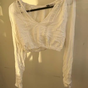 Vit croppad blus med spetsdetaljer - Säljer en vit croppad blus från H&M med långa ärmar och v-ringning. Blusen har fina spetsdetaljer framtill och är gjord i ett lätt bomullstyg. Originalet är den en ganska lång tröja det är jag som har sytt om den till en croppad blus.