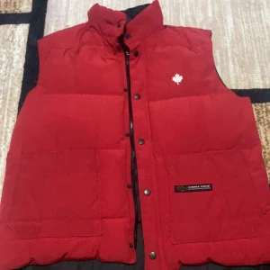 Röd dunväst från Canada Goose - Snygg röd dunväst från Canada Goose med vit lönnblad-logga på bröstet. Västen har hög krage, två stora fickor framtill och stängs med både dragkedja och tryckknappar. Insidan är svart och quiltad för extra värme. Perfekt för kalla dagar.