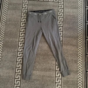 Grå mjukisbyxor Calvin Klein Jeans M - Grå mjukisbyxor från Calvin Klein Jeans med elastisk midja och snörning. Byxorna har smal passform, muddar vid benslut och tydlig logga på midjebandet. Perfekta för chill eller träning, tillverkade i mjukt bomullsmaterial.