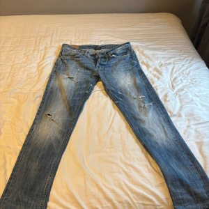 Blå slitna jeans från Dondup - Säljer ett par blå jeans från Dondup med snygga slitningar och distressed detaljer på benen. Modellen har klassisk femficksdesign, rak passform och är tillverkad i 100% bomull. Perfekt för dig som gillar en cool och avslappnad stil.