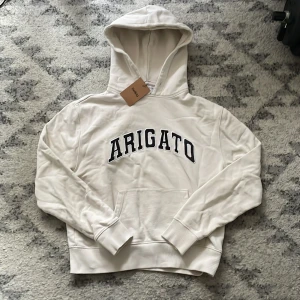 Vit hoodie från Axel Arigato - Snygg vit hoodie från Axel Arigato med svart broderad text på bröstet. Klassisk känguruficka framtill, ribbade muddar och huva. Tillverkad i mjuk bomullsfrotté för extra komfort. Säljer tröjan för att den inte passade mig efter jag köpte den från Sellpy. Tröjan är k fint skick utan några skador.