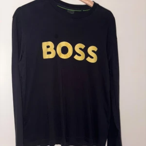 Svart långärmad BOSS-tröja i bomull - Svart långärmad tröja från BOSS med stor guldfärgad BOSS-logga på bröstet. Tröjan har rund halsringning och är tillverkad i mjuk bomull, perfekt för en clean och stilren look.