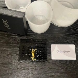 Svart YSL korthållare i skinn - Exklusiv svart korthållare från Yves Saint Laurent i glansigt krokopräglat skinn. Framsidan pryds av den ikoniska YSL-loggan i guld. Perfekt för kort och ID, smidig att ha i fickan eller väskan. Kommer med originalask och äkthetskort.