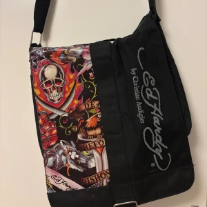 Ed Hardy axelväska  - Svart axel / cross-body väska från Ed hardy. Väskan har justerbar axelrem och rymmer mycket ( t.ex dator, kläder mm). Har några små defekter, men märks inte av och skapar inga problem. Skriv om du har frågor🌸