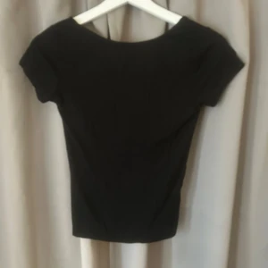 Svart topp med rosett i ryggen - Snygg svart topp med korta ärmar och djup rygg med en stor satinrosett som detalj. Oanvänd och super snygg! 