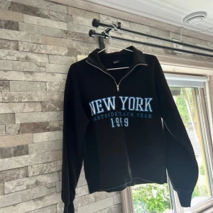 Svart half zip tröja Gina Tricot - Svart långärmad tröja från Gina Tricot med blå och vit text 'NEW YORK' framtill. Tröjan har en halv dragkedja vid halsen och är i mjukt bomullsmaterial. Perfekt för en chill och sportig look.