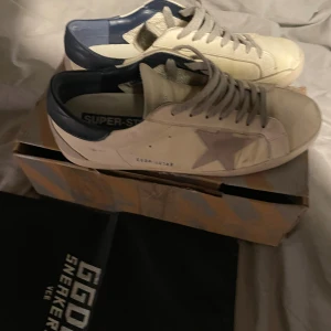 Golden Goose Superstar sneakers vit/blå - Golden Goose Superstar sneakers i vitt skinn med blå häl och grå mockadetaljer. Klassisk stjärna på sidan, grå snörning och platt sula. Snyggt slitna detaljer för en cool look. Kommer med originalkartong och dustbag. Skorna passar 43-44