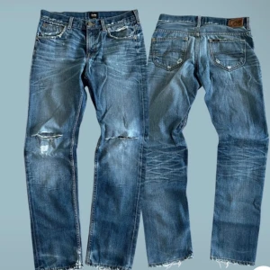 Blå slitna jeans från Lee - Säljer ett par blå jeans från Lee med snygga slitningar och hål vid knäna. Jeansen har klassisk femficksdesign, normal passform och råa, fransiga benslut. Materialet är robust denim i bomull och färgen är tvättad blå med ljusare partier. På lappen står det att jeansen är 27/32 men jag skulle säga att det är mer 29/32