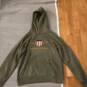 Olivgrön hoodie från GANT - Olivgrön hoodie från GANT med broderad logga och amerikanskt sköldmärke på bröstet. Klassisk känguruficka framtill, huva och ribbade muddar. Perfekt för chill dagar och snygg streetstyle. Materialet är mjuk bomullsmix.