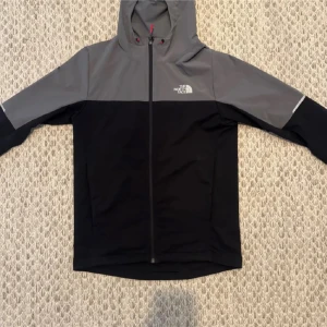 Svart och grå vindjacka från The North Face - Snygg svart och grå vindjacka från The North Face med huva och hel dragkedja. Säljer för att jag aldrig använt den och den är i helt nytt skick. Pris 700kr.
