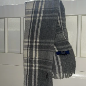 Grå rutig halsduk från Polo Ralph Lauren - Snygg grå halsduk från Polo Ralph Lauren med rutigt mönster i vitt och mörkgrått. Halsduken har klassiska fransar i ändarna och broderad logga. Perfekt accessoar för kyliga dagar och stilren look.