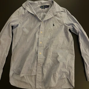 Blå slim fit skjorta från Ralph Lauren - Klassisk ljusblå skjorta från Ralph Lauren i slim fit-modell. Skjortan har långa ärmar, knappar framtill och den ikoniska broderade polospelaren på bröstet. Tillverkad i mjuk bomull och passar perfekt till en clean och stilren look.