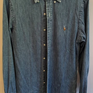 Ralph Lauren jeansskjorta - Snygg blå jeansskjorta från Ralph Lauren. Skjortan är aldrig använd och alltså i nytt skick. Jag säljer den eftersom den tyvärr inte passar mig. Ralph lauren-logot på bröstet. Materialet är mjukt jeans och ärmarna är långärmade med knapp vid manschetten. Perfekt för dig som gillar stilren denim.