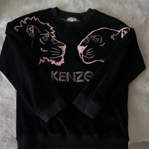 Svart Kenzo sweatshirt med rosa detaljer - Cool svart sweatshirt från Kenzo med broderade rosa lejon- och pantermotiv framtill. Tröjan har rund hals, långa ärmar och ribbade muddar. Mjuk och skön, perfekt för dig som vill sticka ut med en unik design. (Barn tröja).